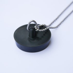 Könnte beinhalten: Ein schwarzer Gummi-Waschbeckenstöpsel mit einer silbernen Metallkette. Der Stöpsel ist rund und hat eine kleine Schlaufe zum Befestigen der Kette. Die Kette besteht aus kleinen, runden Metallperlen.