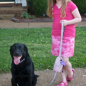 May include: A young girl with long blonde hair wearing a pink shirt and a pink and white tie-dye skirt walks a black labrador retriever on a leash. The girl is smiling and holding the leash in her right hand. The dog is looking at the camera.