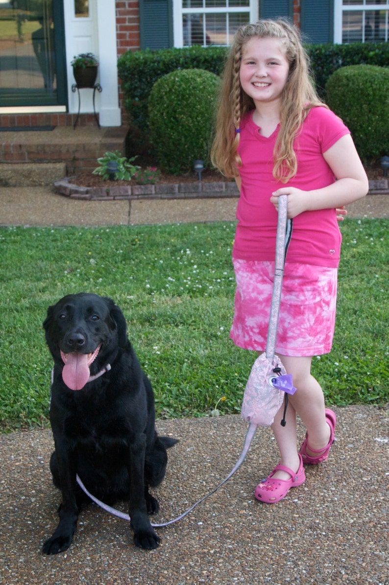 May include: A young girl with long blonde hair wearing a pink shirt and a pink and white tie-dye skirt walks a black labrador retriever on a leash. The girl is smiling and holding the leash in her right hand. The dog is looking at the camera.