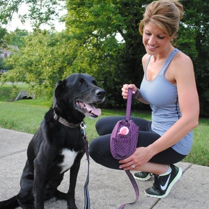 May include: A woman in a blue tank top and black pants walks with a black dog on a leash. She is holding a purple and black patterned bag.