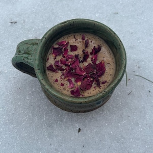 May include: A green ceramic mug filled with a brown beverage topped with dried rose petals. The mug is sitting on a white snowy surface.