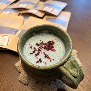 May include: A blue and green ceramic mug with a handle, filled with a blue beverage topped with red dried hibiscus flowers. The mug is sitting on a crocheted coaster.