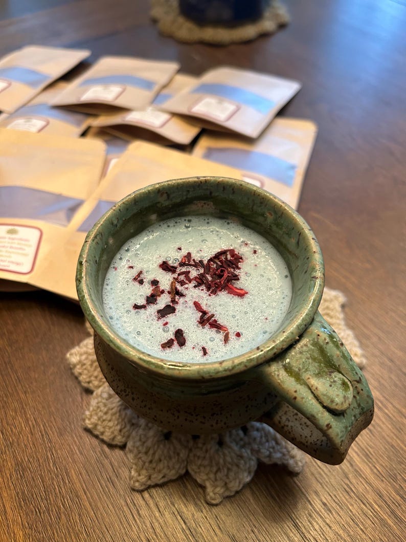 Puede incluir: Una taza de cer&aacute;mica azul y verde con asa, llena de una bebida azul cubierta con flores de hibisco secas rojas. La taza est&aacute; sobre un posavasos de ganchillo.
