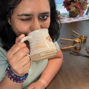 Puede incluir: Una persona est&aacute; bebiendo de una taza de cer&aacute;mica marr&oacute;n y blanca moteada. La taza tiene un asa y una forma redondeada. La persona lleva una camisa verde claro y una pulsera de cuentas azules y moradas.