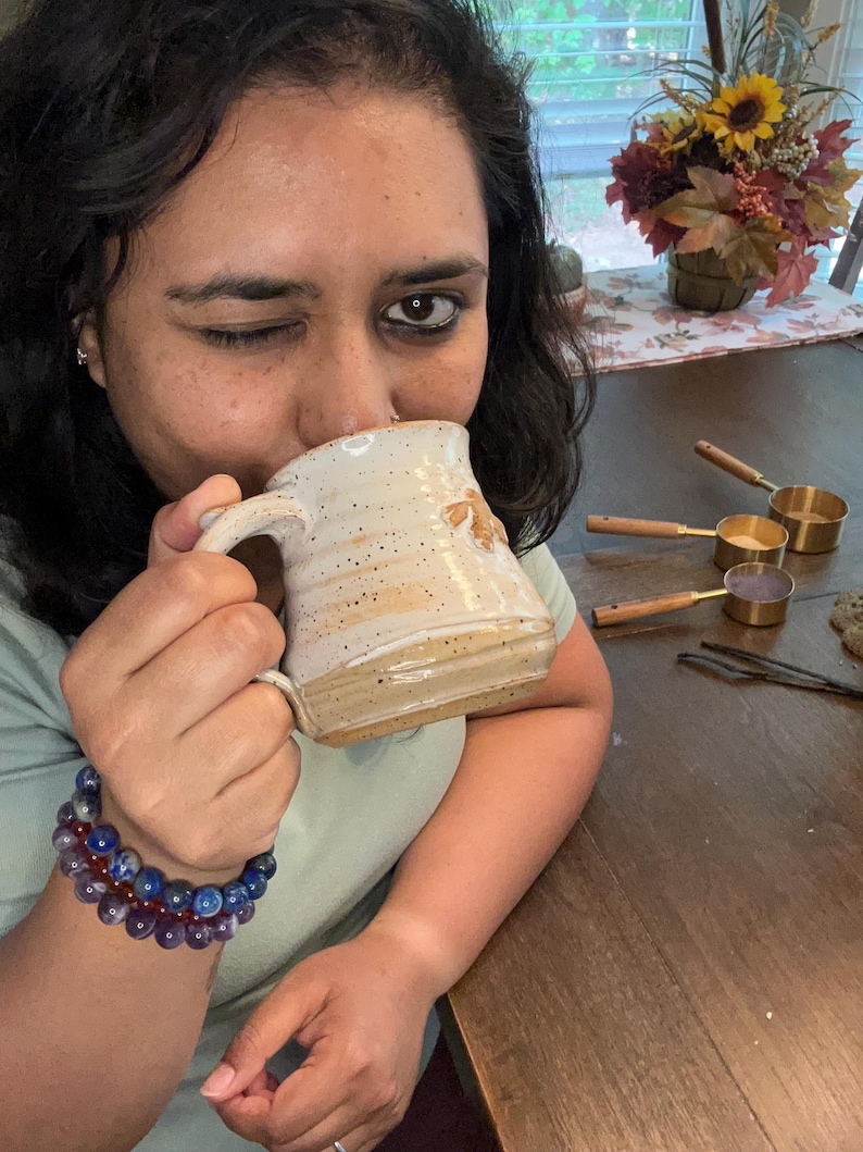 Puede incluir: Una persona est&aacute; bebiendo de una taza de cer&aacute;mica marr&oacute;n y blanca moteada. La taza tiene un asa y una forma redondeada. La persona lleva una camisa verde claro y una pulsera de cuentas azules y moradas.