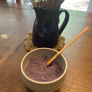 Puede incluir: Un peque&ntilde;o taz&oacute;n blanco lleno de polvo morado, una cuchara de madera descansando en el borde del taz&oacute;n. Una jarra azul con tallos de lavanda secos se encuentra sobre un posavasos de ganchillo en una mesa de madera.