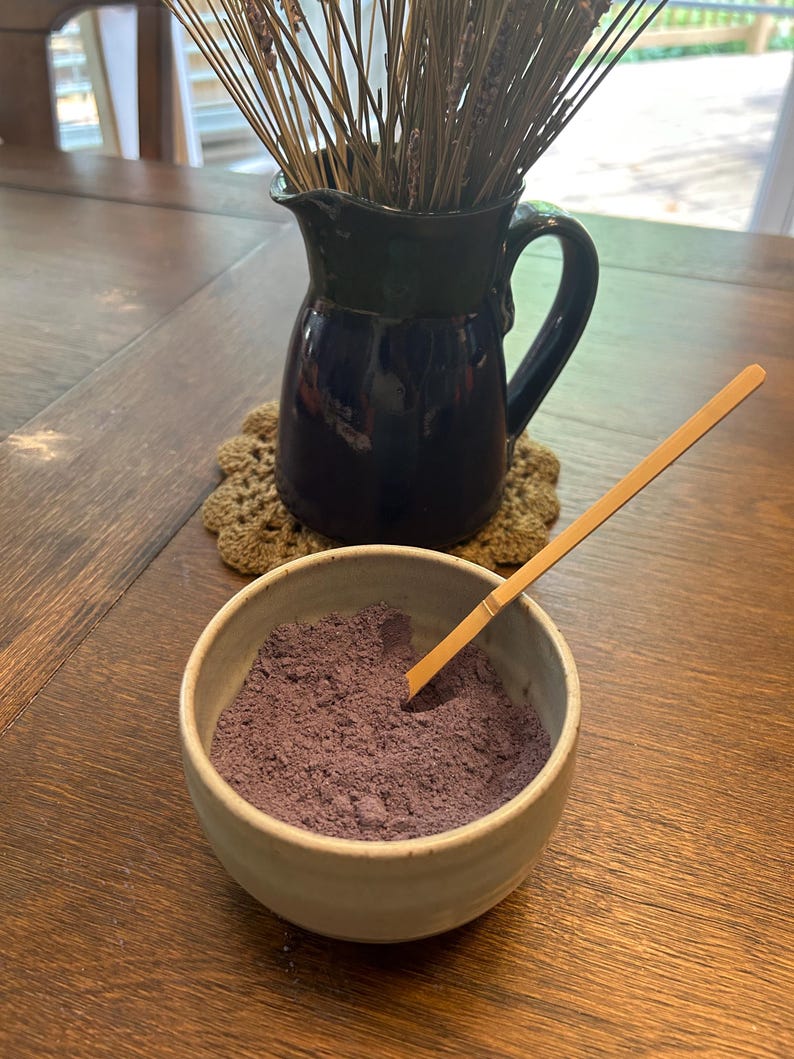 Puede incluir: Un peque&ntilde;o taz&oacute;n blanco lleno de polvo morado, una cuchara de madera descansando en el borde del taz&oacute;n. Una jarra azul con tallos de lavanda secos se encuentra sobre un posavasos de ganchillo en una mesa de madera.