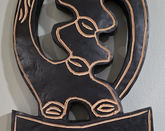 May include: A black wooden wall hanging with a carved design. The artwork features a central shape with a series of interconnected forms, and a decorative border. The design is accented with light brown details.