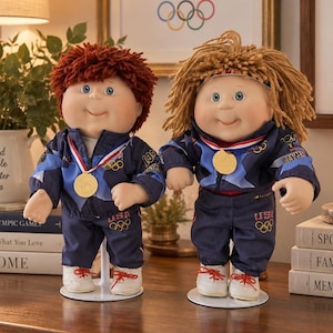 May include: Two dolls with yarn hair wearing navy blue USA Olympic tracksuits and white sneakers. Each doll has a gold medal around its neck. The doll on the left has short reddish-brown hair, and the doll on the right has longer light brown hair.