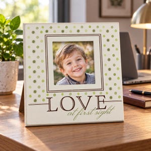 May include: A square cream-colored picture frame featuring a light green polka dot pattern and the dark brown text "LOVE at first sight" printed at the bottom. The frame contains a photograph of a smiling child with light brown hair.