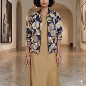 May include: A navy blue bomber jacket featuring a beige and gold chain-link and floral pattern, worn over a solid mustard yellow sleeveless dress. The jacket has a ribbed collar, cuffs, and hem, with a central zipper closure and long sleeves.