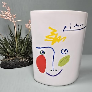 May include: White ceramic vase with a colorful abstract face design. The face features a red cheek, green and blue accents, and the name "Picasso" in blue. A small succulent plant is to the left of the vase.