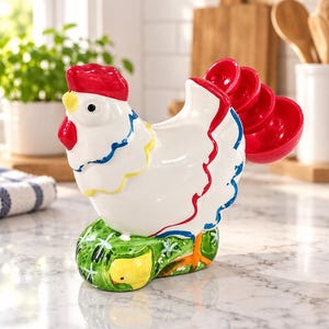 May include: A white ceramic chicken-shaped measuring cup set with a red comb and wattle, blue and yellow neck accents, and a green base featuring a small yellow chick. The tail feathers consist of stacked red measuring cups.