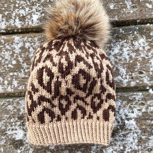 May include: A knitted winter hat with a brown and tan leopard print pattern. The hat features a fluffy, light brown pom-pom on top. The brim is a solid tan color.
