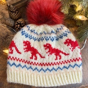 Puede incluir: Gorro de punto blanco con un pompón rojo y un patrón de dinosaurio rojo, blanco y azul.