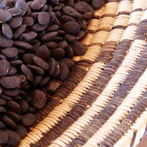 Peut inclure: Gros plan sur des graines sombres et ovales débordant d'un panier tressé. Le panier présente un motif de bandes tissées beiges, noires et blanches alternées, créant une surface texturée. Les graines sont d'un noir profond et uniforme.