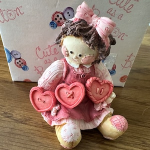 May include: A pink and brown rag doll figurine holding two red heart-shaped buttons. The doll is sitting on a wooden surface. The doll has a pink bow in her hair and a pink dress with white polka dots. The doll is part of a collection called "Cute as a Button".