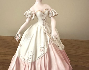 May include: A porcelain figurine of a woman in a white and pink dress with gold accents. She is wearing a lace-trimmed bodice and has a flower in her hair.