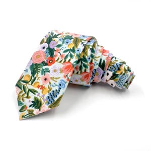 May include: A necktie featuring a light blue background with a dense, colorful floral pattern. The design includes various flowers in shades of pink, orange, yellow, and purple, interspersed with green leaves and small blue berry-like clusters.