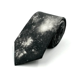 May include: A necktie featuring a black and white galaxy print with scattered white star-like clusters against a dark charcoal background. The fabric has a textured appearance, and the tie is rolled into a coil, showing the pointed end.