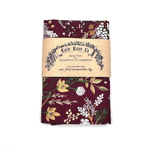 May include: A folded burgundy bow tie with a floral pattern of white berries and gold and green leaves. A brown paper band wraps around the center, displaying the text "Kate Rose Co. BOW TIES & NECKWEAR ACCESSORIES -HANDMADE- est. 2013 Louisville, Ky."