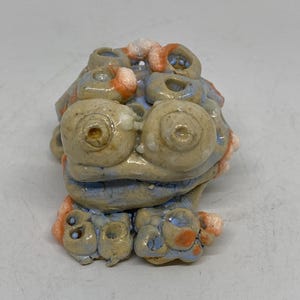 May include: A ceramic sculpture of a creature with large, round eyes and a textured surface. The sculpture features a color palette of beige, blue, and orange, with a whimsical and organic design. The sculpture is a unique decorative item.