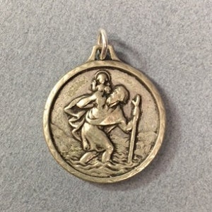 May include: A silver-toned pendant featuring a detailed image of Saint Christopher carrying a child. The pendant is circular and has a smooth, polished finish.