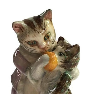 May include: A porcelain figurine of two cats, one brown and white with a purple dress, the other brown and white with a yellow and orange fruit in its paws.
