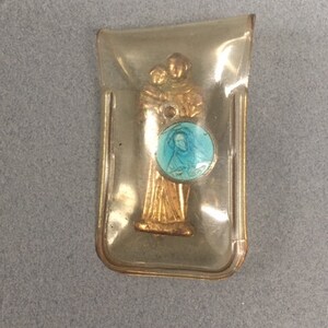 May include: A gold-colored religious figurine of a saint holding a child, encased in a clear plastic case. The saint is wearing a blue robe with a gold trim and a halo. The child is wearing a white robe. The saint is holding a round blue and white medallion with a picture of a person on it.
