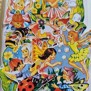 May include: A collection of vintage fairy and insect cutouts. The fairies have yellow, orange, and blue wings and dresses. A ladybug and a bumblebee are also present. The cutouts are on a white background. A ruler is in the image, with measurements in centimetres.