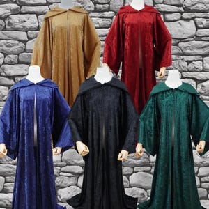 May include: Five velvet hooded cloaks in various colors: gold, red, blue, black, and green. Each cloak has a long, flowing design with wide sleeves and a front zipper. The cloaks are displayed on mannequins against a stone wall background.