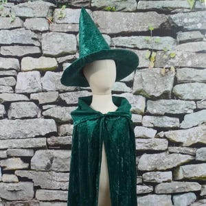 May include: A green velvet witch costume set, including a pointed hat with a wide brim and a hooded cape. The costume is displayed on a mannequin against a stone wall backdrop.
