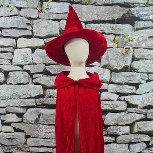 May include: A red witch hat with a wide brim and a matching red cape. The hat is tall and pointed, and the cape has a hood. The costume is displayed on a mannequin against a stone wall background.