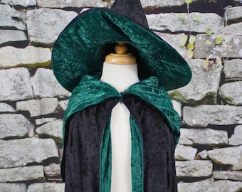 May include: A black and green witch costume set. The set includes a pointed black hat with a wide brim and a black cape with green trim. The costume is displayed on a mannequin against a stone wall background.