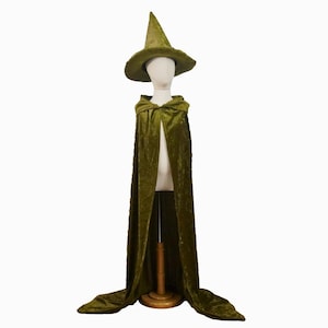 May include: A green velvet witch costume set on a mannequin. The set includes a pointed hat and a hooded cape. The cape is floor-length and has a flowing design. The hat has a wide brim and a textured appearance. The costume is displayed on a wooden stand.