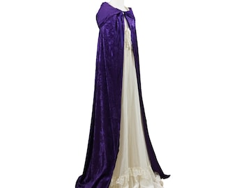 May include: A long purple velvet hooded cape with a gathered neckline. The cape is worn over a white dress with lace trim.