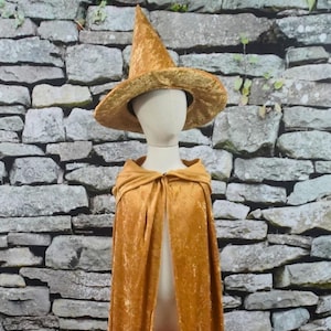 May include: A golden-yellow witch costume set, including a pointed hat and a hooded cape. The hat has a wide brim and the cape drapes down. The costume is displayed on a mannequin against a stone wall background.