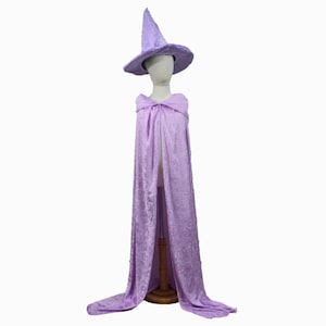 May include: A lavender witch costume set, including a pointed hat and a long cape. The cape is draped over a mannequin, showcasing the flowing fabric and design. The costume is designed for dress-up or Halloween.