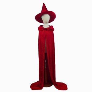 May include: A red witch costume set, including a pointed hat and a long cape. The hat has a wide brim and a conical top. The cape is floor-length and has a hood. The costume is displayed on a mannequin.