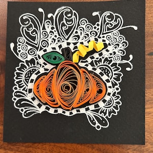 Op de afbeelding: Een handgemaakt papier quilling kunstwerk met een oranje pompoen met een groen blad en een gele stengel, tegen een zwarte achtergrond. De pompoen is omgeven door witte decoratieve krullen en patronen, wat een feestelijk herfstthema creëert.