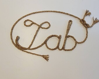 May include: A decorative sign spelling "Lab" made from natural rope. The rope is looped to form the letters and has frayed ends. The sign is designed to be hung and is displayed against a white background.