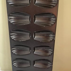 Può includere: Una teglia per madeleine marrone scuro con 12 stampi a forma di conchiglia. La teglia è rettangolare con un foro nella parte superiore per appenderla. Gli stampi hanno un design a coste, perfetto per cuocere le classiche madeleine francesi.