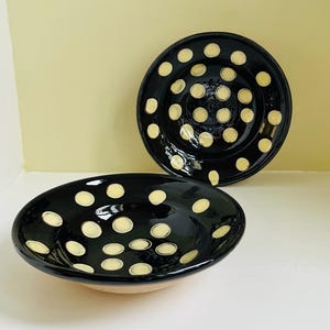 May include: Two black ceramic bowls with a glossy finish. Each bowl is decorated with evenly spaced, cream-colored polka dots. The bowls are of different sizes and are displayed on a white surface.