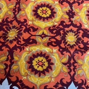May include: Close-up of a fabric with a repeating floral pattern. The design features large, stylized flowers in shades of yellow, orange, and burgundy. The background is a lighter orange, and the fabric has a textured appearance. The bottom edge has a scalloped border.