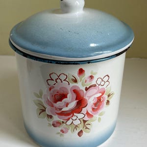Peut inclure: Une boîte en émail blanche avec un bord et un couvercle bleus, ornée d'un motif floral de roses roses et rouges. La boîte a un petit bouton blanc sur le couvercle.