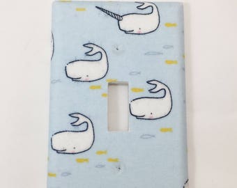 May include: Light blue light switch cover with a white narwhal and whale pattern. The narwhals have a yellow horn and the whales have yellow fish swimming around them.