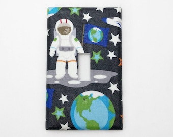 May include: A decorative light switch cover featuring a space theme. The cover has a dark gray background with white stars, planets, and an astronaut. The astronaut is holding a flag with a globe on it.