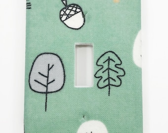 May include: A light green light switch cover with a white, gray, and gold woodland pattern. The pattern includes trees, acorns, and leaves.