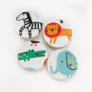 May include: Four round fabric buttons with different animal designs. The buttons feature a zebra, a lion, a crocodile, and an elephant. The buttons are white with colorful animal illustrations.