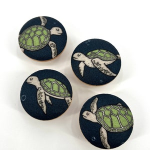 May include: Four round fabric buttons with a dark blue background and a green sea turtle design. The turtles are swimming in a light blue ocean with white bubbles.